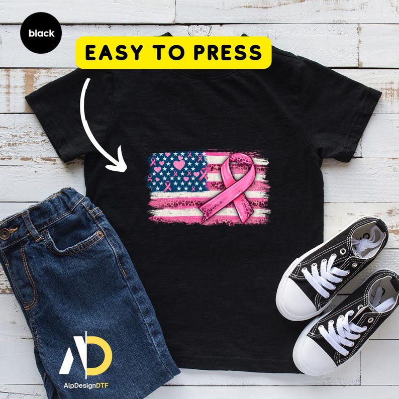 May include: Black t-shirt featuring a pink ribbon and an American flag design with hearts and stars. The words "EASY TO PRESS" are in a yellow rectangle above the design. A pair of black and white sneakers and blue jeans are next to the shirt.