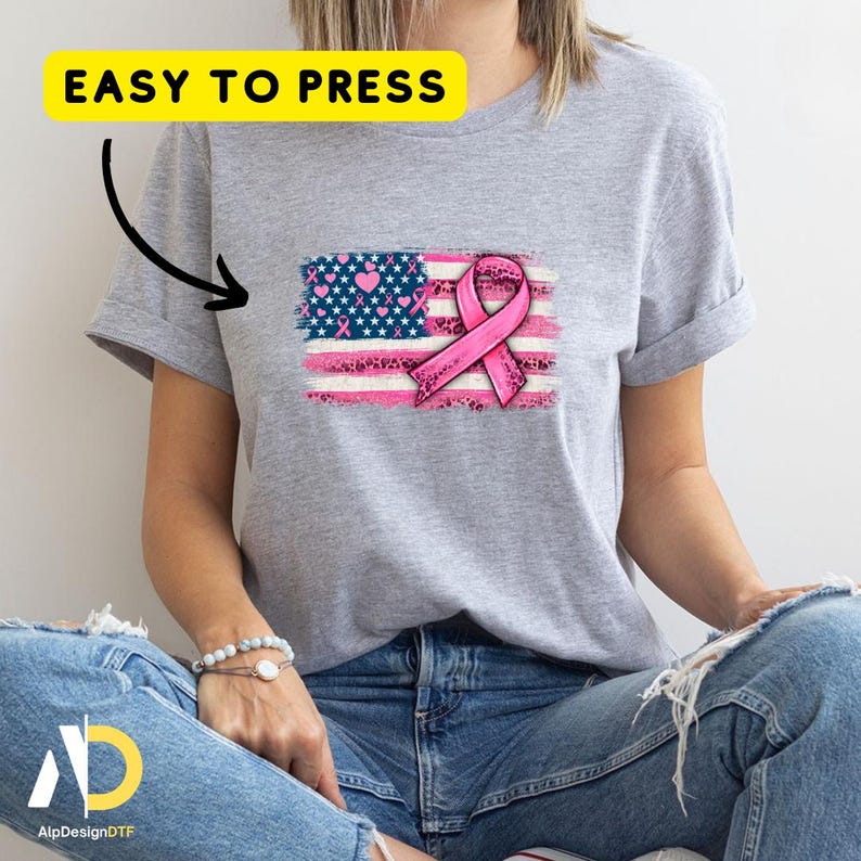May include: A gray t-shirt with a graphic of a pink ribbon and an American flag design. The flag incorporates pink ribbons, hearts, and stars. The text "EASY TO PRESS" is in a yellow rectangle above the design.