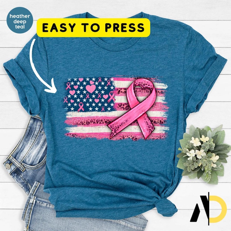 May include: Teal t-shirt featuring a distressed American flag design with pink stripes and a pink ribbon, symbolizing breast cancer awareness. The flag incorporates pink hearts and ribbons. The text "EASY TO PRESS" is displayed in a yellow rectangle.
