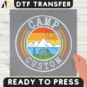May include: DTF TRANSFER ready to press. A circular design with the words "CAMP CUSTOM" encircling a landscape scene. The scene includes mountains, a lake, a tent, and a sun. The background is a gradient of orange and yellow.