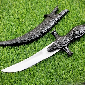 May include: A black decorative dagger with a silver blade and intricate floral patterns. The dagger is on a green grassy surface.