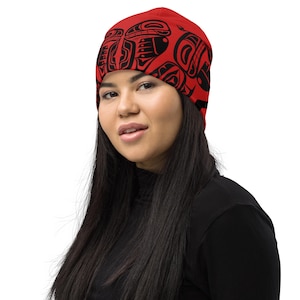 Puede incluir: Un gorro rojo con un diseño tribal negro. El gorro está hecho de un material suave y lo lleva una persona que lleva un cuello alto negro.