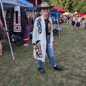 May include: A long, white, open-front kimono-style jacket featuring bold black and red formline graphic designs. The garment is worn over a black top and blue distressed denim jeans, paired with a tan wide-brimmed hat and black ankle boots.