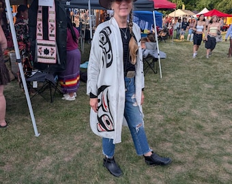 Może przedstawiać: Długa, biała, rozpinana z przodu kurtka w stylu kimono z wyrazistymi czarnymi i czerwonymi wzorami graficznymi. Ubiór noszony jest na czarny top i niebieskie dżinsy z przetarciami, w połączeniu z beżowym kapeluszem z szerokim rondem i botkami.