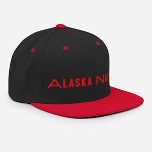 May include: A black and red baseball cap with the embroidered text "ALASKA NAT". The cap has a flat red peak and red eyelets. The top button is also red.