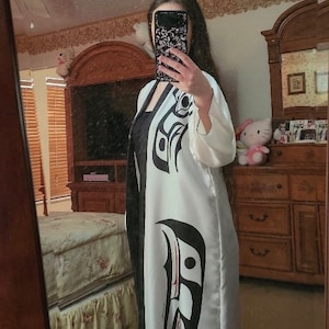 May include: A white and black patterned robe with a Native American design. The robe has long sleeves and a flowing silhouette. The design features bold black shapes on a white background.