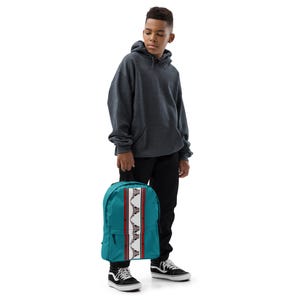 May include: Teal backpack with a central vertical design featuring a white stripe flanked by red stripes and a repeating pattern. The backpack is held by a person wearing a gray hoodie and black pants.