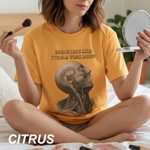May include: Citrus yellow t-shirt with the text "DOES IT LOOK LIKE I'VE HAD WORK DONE?" above a detailed anatomical illustration of a human head and neck. The shirt is a solid color and has a crew neck.