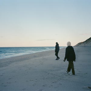 Puede incluir: Dos personas caminan por una playa arenosa cerca del océano. El cielo es azul pálido y el agua es azul más oscuro. Una persona lleva una chaqueta negra, un gorro blanco y pantalones verde oliva. La otra persona lleva una chaqueta oscura.