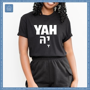 May include: Black crew neck t-shirt with the word "YAH" in large white block letters, with Hebrew characters and the letter "T" below. The shirt is part of a clothing collection.