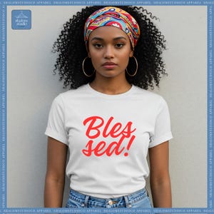 Puede incluir: Camiseta blanca con el texto rojo "Blessed!". Una mujer con cabello negro rizado lleva la camiseta, pendientes de aro dorados y un pañuelo colorido. La camiseta tiene las mangas dobladas. El fondo es una pared gris claro.
