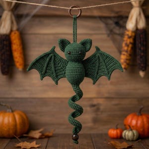 May include: A dark green crocheted bat ornament featuring pointed ears, wide wings, and a long, spiraling tail. The bat is suspended from a brown wooden ring at the top and finished with a small, dark brown wooden bead at the bottom tip.