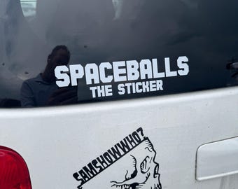 May include: White sticker on a car window with the text "SPACEBALLS THE STICKER" in a bold, white font. Below is a black and white skull graphic with the text "SAMSHOWWHO?" in a stylized font.