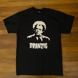 May include: Black t-shirt with a white silhouette of a man with an afro wearing glasses and a suit. The text "DRANZIG" is printed below the silhouette.