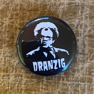 May include: Black and white button with a silhouette of a person with a large afro and the word "Dranzig" printed in white.