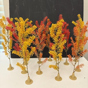 May include: Artificial floral stems in yellow, orange, and red hues. Each stem is set in a small, beige base. The stems are arranged on a white surface, with a dark background.