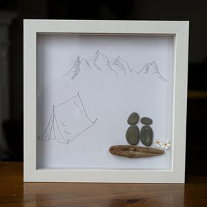 Op de afbeelding: Een ingelijst kunstwerk van kiezelsteen dat een stel voorstelt dat op een boomstam zit voor een tent en bergen. De bergen en de tent zijn getekend met zwarte inkt op wit papier. Het stel is gemaakt van grijze kiezelsteen en er zijn witte bloemen op de boomstam.