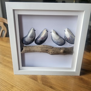 Può includere: Un'immagine incorniciata bianca con quattro piccoli uccelli grigi e bianchi appollaiati su un ramo. Gli uccelli sono fatti di pietre dipinte.