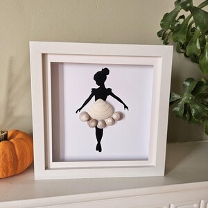 May include: A framed artwork featuring a black silhouette of a ballerina. The ballerina's tutu is crafted from seashells, and the artwork is encased in a white frame. The background is white, and the frame is displayed on a white surface.