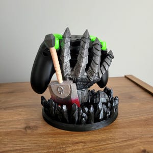May include: A black video game controller stand featuring a sculpted metallic silver gauntlet hand holding a large cleaver with a tan handle and red blood-like paint. The base is decorated with black jagged crystal-like formations.