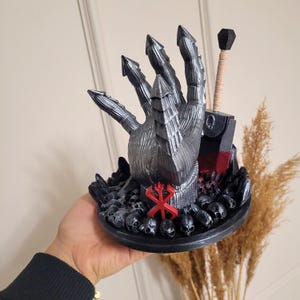 May include: A decorative sculpture featuring a metallic silver clawed hand rising from a base of small black skulls. A red symbol is centered on the wrist, and a sword with a tan handle and a blood-stained blade stands upright behind the hand.