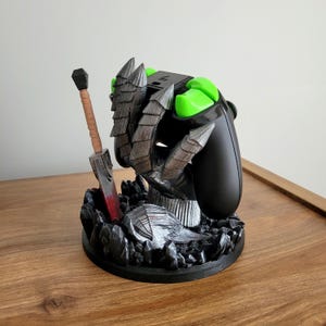 May include: A black video game controller stand featuring a sculpted grey monster claw gripping the device. The base is decorated with dark rocky textures and a miniature sword with a tan handle and a blade featuring a red accent at the base.