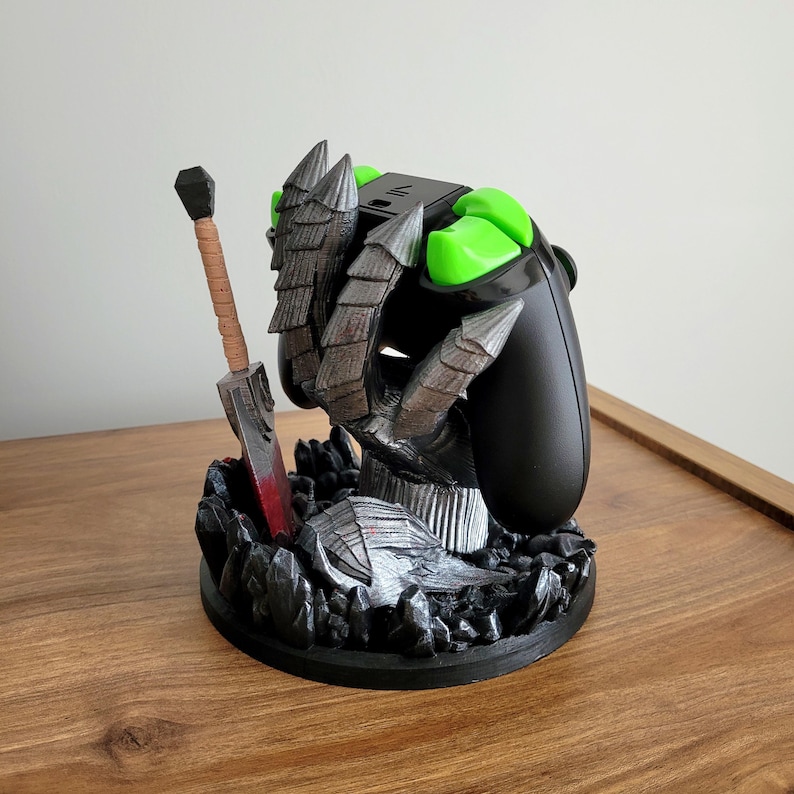 May include: A black video game controller stand featuring a sculpted grey monster claw gripping the device. The base is decorated with dark rocky textures and a miniature sword with a tan handle and a blade featuring a red accent at the base.