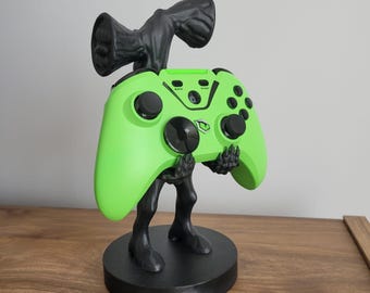 May include: A black and green Siren Head-themed controller stand. The stand features a green controller held by black hands and legs, with a Siren Head figure on top. The base is black and round.