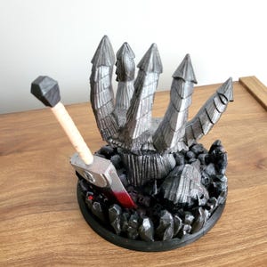 May include: A 3D printed figurine featuring a silver, multi-pointed claw-like structure emerging from a dark, rocky base. A sword with a light-colored handle and a blade marked with a red stain is embedded into the base among the dark stones.