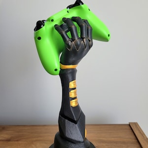 May include: A black and gold robotic hand-shaped gaming controller stand holding a bright green controller. The stand has gold accents and a detailed, mechanical design, perfect for displaying gaming accessories.
