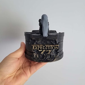 May include: A black, cylindrical decorative object with the text "EXPEDITION 33" in gold lettering. A small figure with long grey hair is on top. The object is held in a hand against a white background.