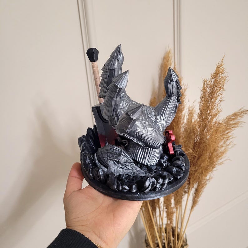 May include: A 3D printed figurine featuring a silver-colored armored hand with layered plating reaching upward, positioned on a circular base of dark rocks. A large sword with a black blade and a tan handle stands upright behind the armored hand.