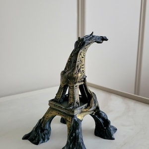 May include: A decorative sculpture of the Eiffel Tower featuring a distorted, melting aesthetic. The structure is finished in a combination of matte black and metallic gold paint, with the base and top appearing to drip or flow downwards.