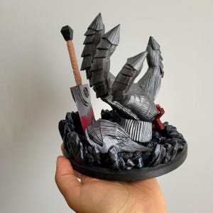 May include: A 3D printed figurine featuring a large, armored, metallic gray clawed hand emerging from a dark, rocky base. Beside the hand stands a tall, rectangular sword with a tan handle and a blade marked with red, blood-like accents.