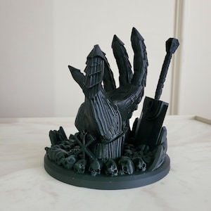 May include: A matte black 3D printed sculpture featuring a large armored hand emerging from a base covered in small skulls. A sword stands upright next to the hand, and a geometric symbol is visible on the base among the skull pile.
