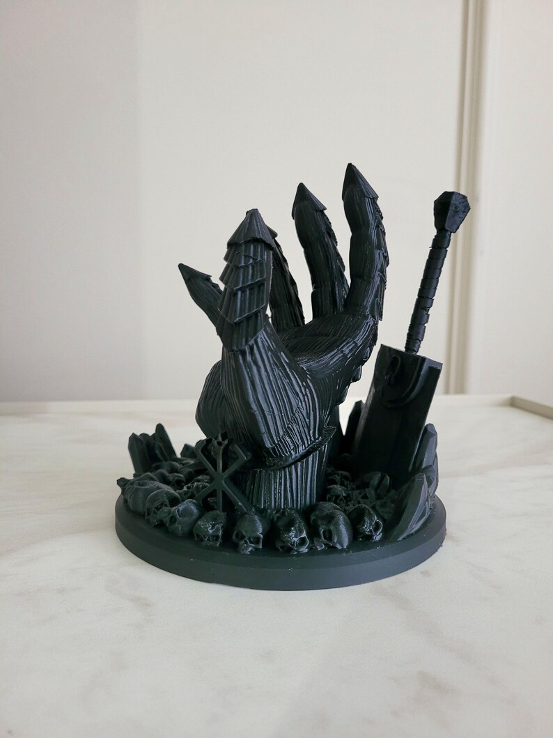 May include: A matte black 3D printed sculpture featuring a large armored hand emerging from a base covered in small skulls. A sword stands upright next to the hand, and a geometric symbol is visible on the base among the skull pile.