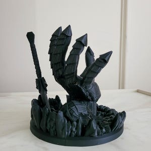 May include: A matte black 3D printed sculpture featuring a large armored hand reaching upward from a base of jagged crystal-like formations, accompanied by a long sword standing upright next to the hand, all set on a circular base.