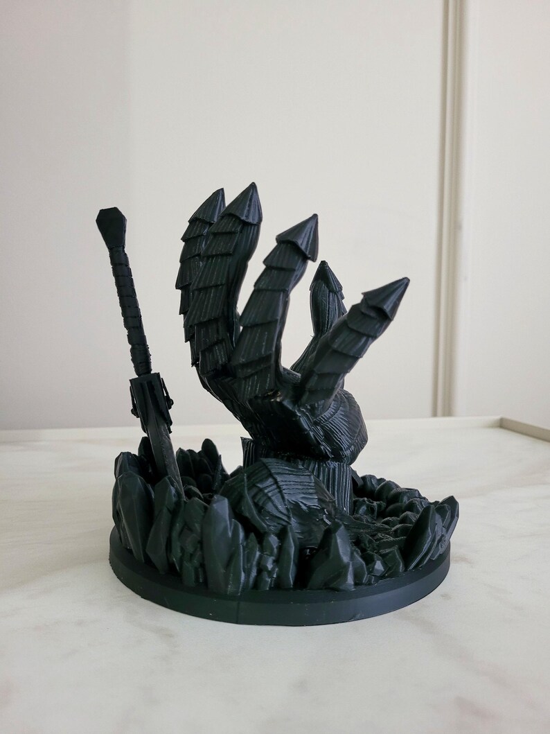 May include: A matte black 3D printed sculpture featuring a large armored hand reaching upward from a base of jagged crystal-like formations, accompanied by a long sword standing upright next to the hand, all set on a circular base.