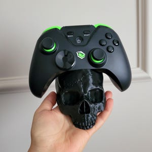 May include: Black gaming controller with green accents, resting on a black skull. The controller has buttons labeled "Back" and "Start". The skull is a decorative item, likely for a gaming setup.
