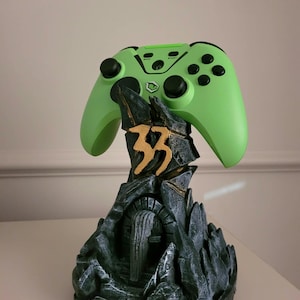 May include: A bright lime green video game controller rests on a dark grey, rock-textured display stand. The stand features a central gold-coloured symbol resembling the number 33 and a sculpted archway at the base with a weathered stone appearance.
