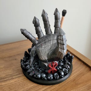 May include: A dark gray, armored gauntlet sculpture featuring five pointed, segmented fingers. The base is adorned with numerous small black skulls and a prominent red symbol. A thin, light-colored rod with a black tip extends from the back.