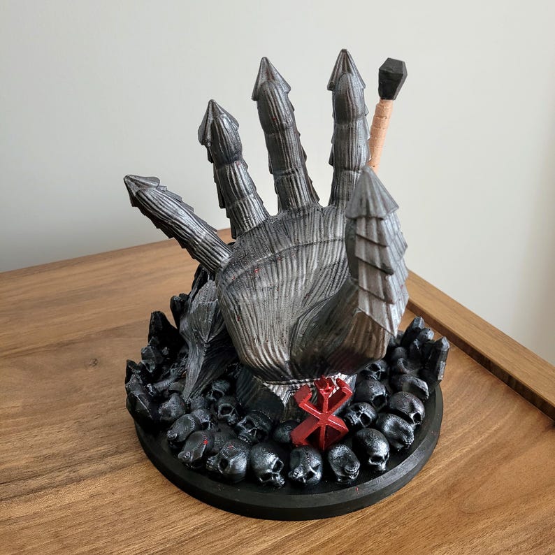 May include: A dark gray, armored gauntlet sculpture featuring five pointed, segmented fingers. The base is adorned with numerous small black skulls and a prominent red symbol. A thin, light-colored rod with a black tip extends from the back.