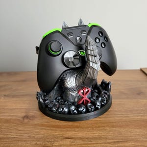 May include: A black video game controller rests on a dark, sculpted stand featuring a metallic silver armored hand reaching upward. The base is decorated with numerous small skulls and a prominent red symbol, providing a sturdy display mount.