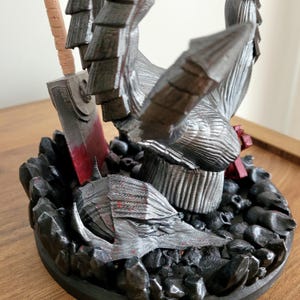 May include: A 3D printed figurine featuring a metallic silver armored hand reaching upward, positioned above a silver helmet with red paint splatters. The base is composed of numerous small black skulls and rocks, with a large sword blade nearby.