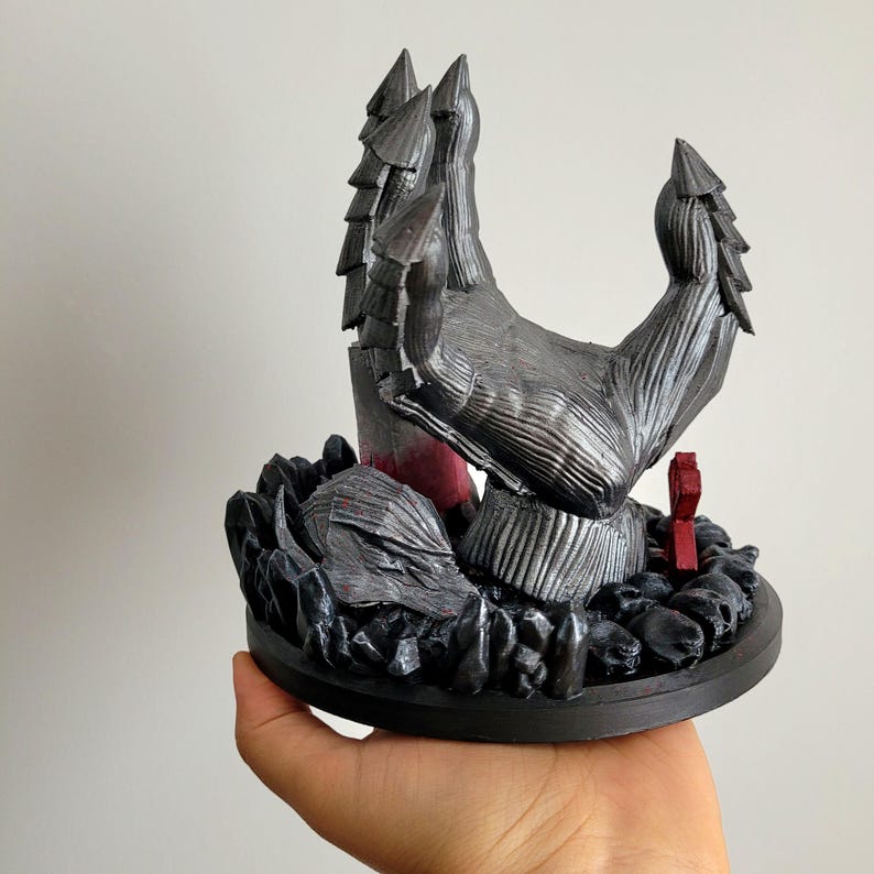 May include: A silver-colored, multi-fingered claw sculpture emerging from a base of dark, jagged rocks and skulls. The piece features a small, upright red rectangular element near the base, with textured, layered details on the claw segments.