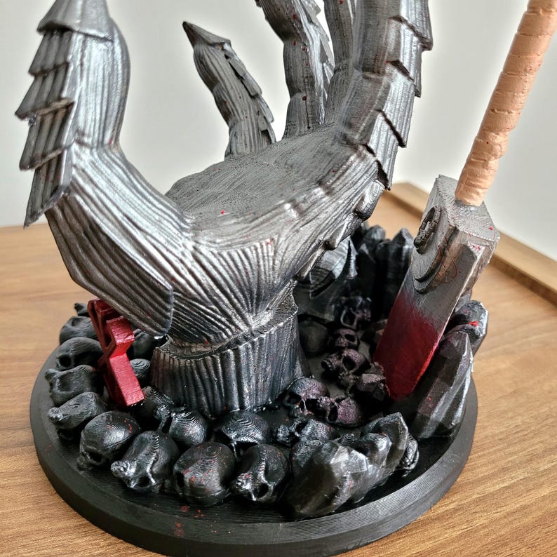 May include: A silver-colored, textured clawed hand sculpture rising from a circular base covered in small black skulls. A red-painted rectangular object and a metallic-looking blade with a tan handle are positioned among the skulls on the base.