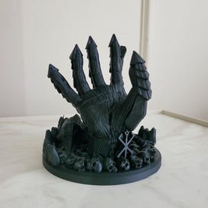 May include: A matte black 3D printed sculpture of a large, armored hand emerging from a base covered in small skulls and jagged rock formations. The hand features segmented, pointed fingers and a prominent brand symbol etched on the wrist area.