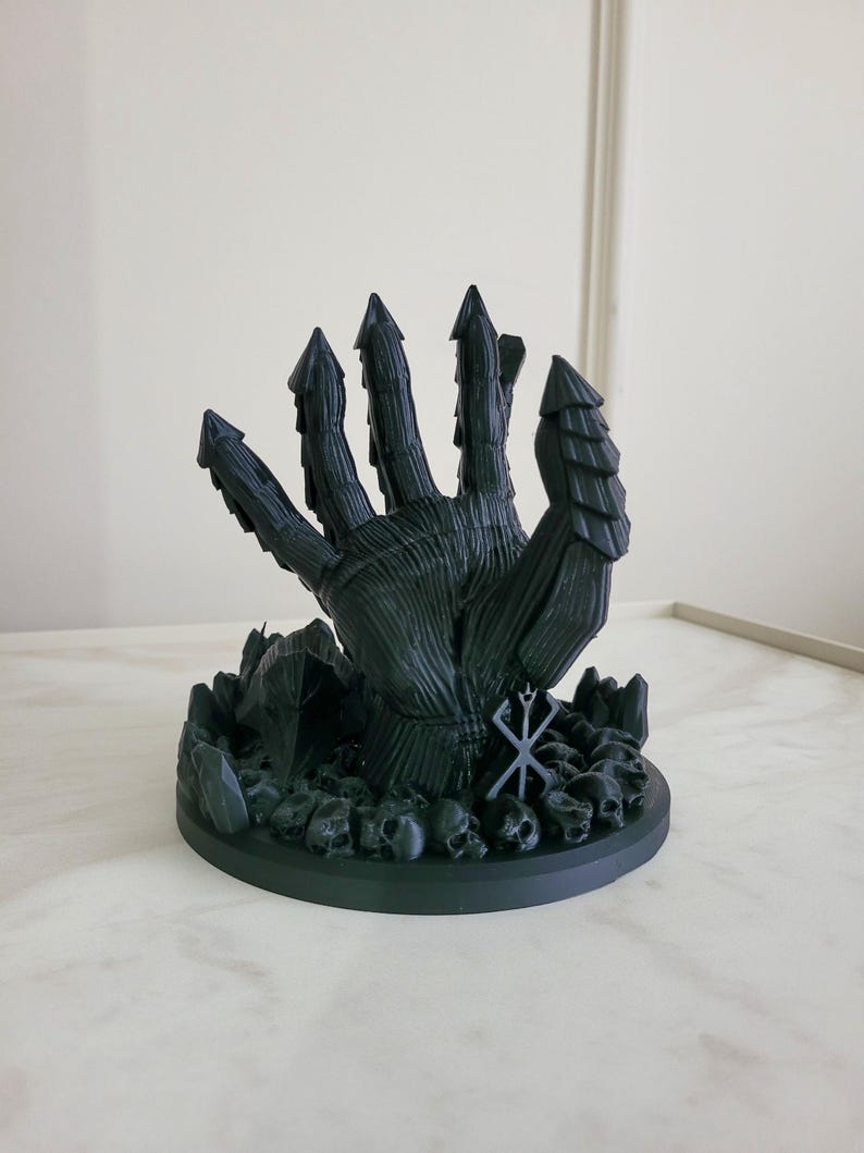 May include: A matte black 3D printed sculpture of a large, armored hand emerging from a base covered in small skulls and jagged rock formations. The hand features segmented, pointed fingers and a prominent brand symbol etched on the wrist area.
