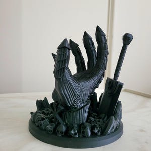 May include: A matte black 3D printed sculpture featuring a large armored hand emerging from a base covered in small skulls. A large sword stands upright next to the hand, and a small cross-like symbol is positioned among the skulls at the base.
