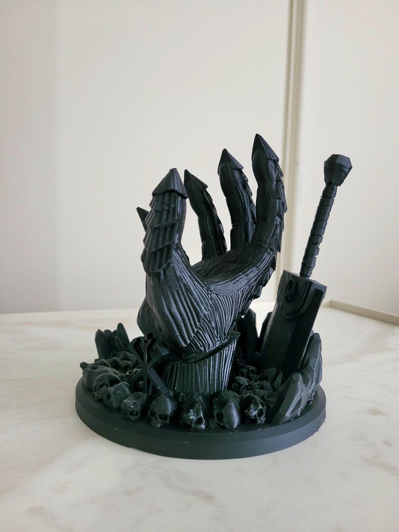 May include: A matte black 3D printed sculpture featuring a large armored hand emerging from a base covered in small skulls. A large sword stands upright next to the hand, and a small cross-like symbol is positioned among the skulls at the base.